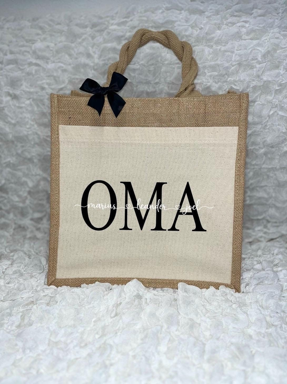 Vor dem weißen Hintergrund befindet sich ein Juteshopper mit einer fast vollflächigen Baumwolltasche darauf. Links auf dem Henkel befindet sich eine schwarze Schleife. Der personalisierte Aufdruck besteht aus Oma & dem/den Wunschnamen.
