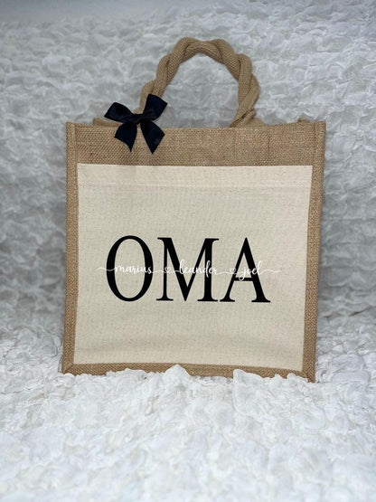 Vor dem weißen Hintergrund befindet sich ein Juteshopper mit einer fast vollflächigen Baumwolltasche darauf. Links auf dem Henkel befindet sich eine schwarze Schleife. Der personalisierte Aufdruck besteht aus Oma & dem/den Wunschnamen.