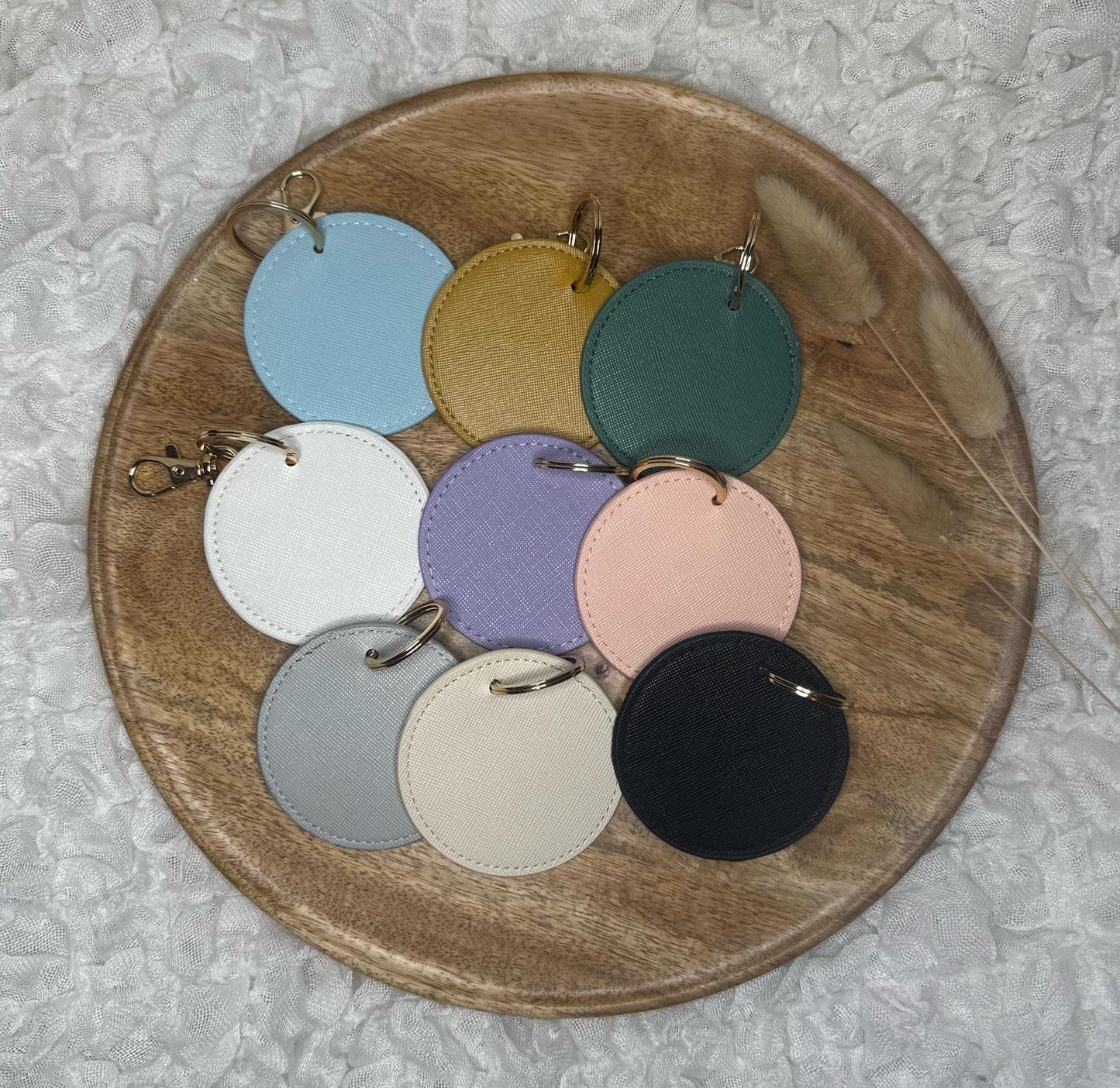 Auf dem weißen Hintergrund befindet sich ein Holztablett. Auf dem Tablett liegen Schlüsselanhänger in verschiedenen Farben mit Anhänger aus glänzendem Metall.