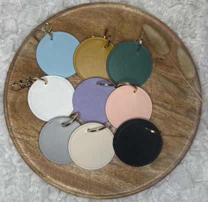 Auf dem weißen Hintergrund liegt ein rundes Holztablett mit Pampasgras. Darauf befinden sich Schlüsselanhänger aus Kunstleder  in verschiedenen Farben. In Grau,Creme, Schwarz, Weiß, Lila, Grün, Sengelb, Babyblau & Rosa.