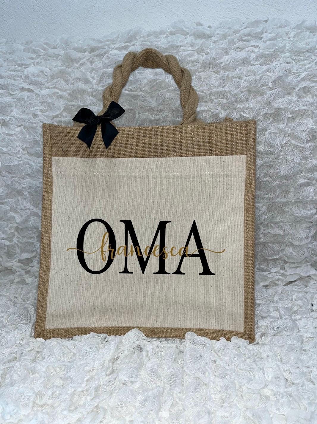 Vor dem weißen Hintergrund befindet sich ein Juteshopper mit einer fast vollflächigen Baumwolltasche darauf. Links auf dem Henkel befindet sich eine schwarze Schleife. Der personalisierte Aufdruck besteht aus Oma & dem/den Wunschnamen.