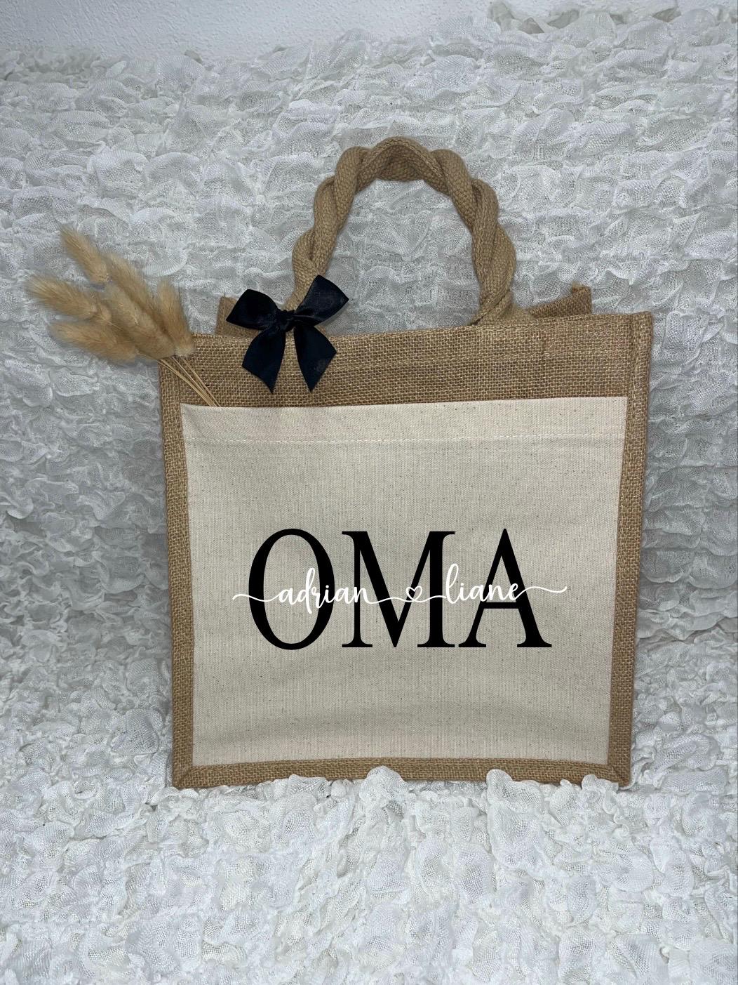 Vor dem weißen Hintergrund befindet sich ein Juteshopper mit einer fast vollflächigen Baumwolltasche darauf. Links auf dem Henkel befindet sich eine schwarze Schleife. Der personalisierte Aufdruck besteht aus Oma & dem/den Wunschnamen.