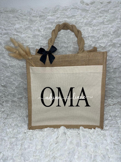 Vor dem weißen Hintergrund befindet sich ein Juteshopper mit einer fast vollflächigen Baumwolltasche darauf. Links auf dem Henkel befindet sich eine schwarze Schleife. Der personalisierte Aufdruck besteht aus Oma & dem/den Wunschnamen.