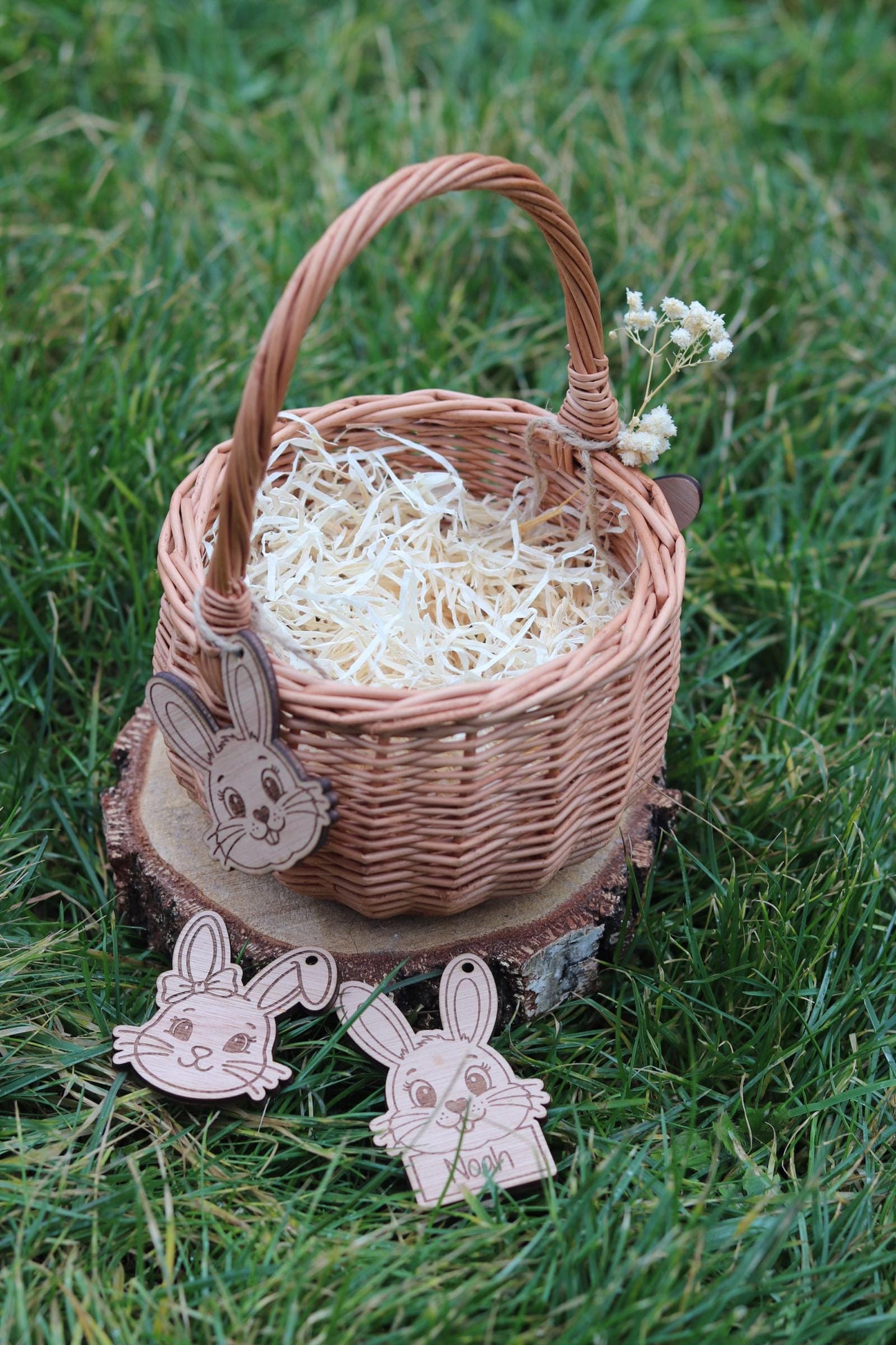 Ein geflochtener brauner Weidenkorb mit Henkel, gefüllt mit Holzwolle, steht auf einer Holzscheibe. Drei Hasen-Ausschnitte aus Holz hängen am Korb und liegen darunter. Ein Zweig weißer Blumen ist am Griff befestigt.